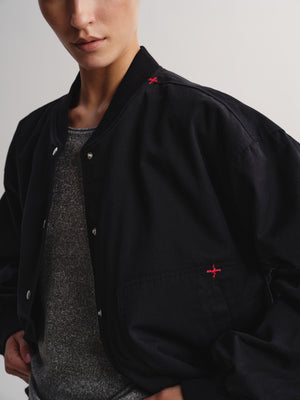 A close-up of a person wearing a black bomber jacket with small red embroidered cross logos on the chest and collar, layered over a gray knit top against a light gray studio background.