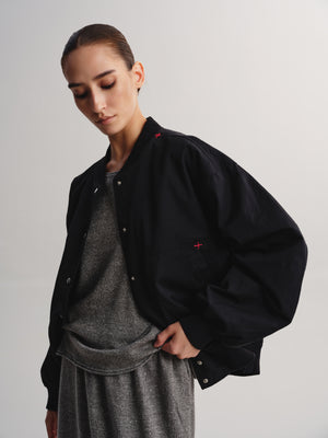 A model wearing a black bomber jacket with small red cross embroidery details on the chest and shoulders, layered over a gray knit top and matching gray skirt, set against a plain light gray studio background.