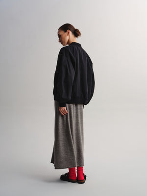 A model is shown from a side-back angle wearing a black bomber jacket, a long gray knit skirt, bright red socks, and black slip-on shoes against a light gray studio background.