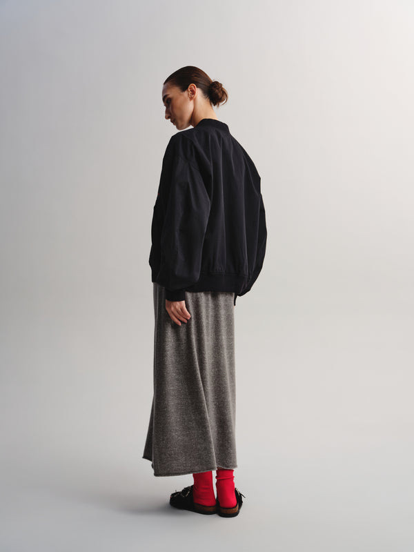 A model is shown from a side-back angle wearing a black bomber jacket, a long gray knit skirt, bright red socks, and black slip-on shoes against a light gray studio background.
