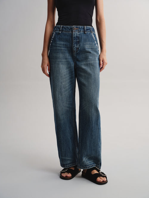 A model wearing a pair of high-waisted, wide-leg blue denim jeans with decorative white embroidery on the pocket edges, styled with a black top and black sandals against a light gray studio background.