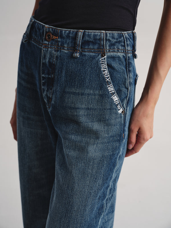 Close-up of a person wearing blue denim jeans with a black top, showing the side pocket detail and front waistband against a plain light background.