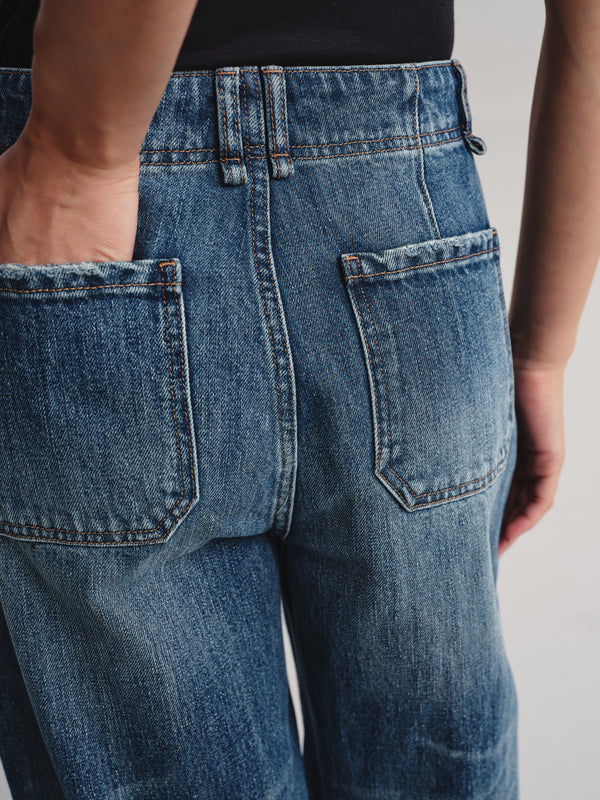 Close-up back view of a person wearing blue denim jeans, showing the back pockets, waistband, and hand in the pocket against a light gray studio background.