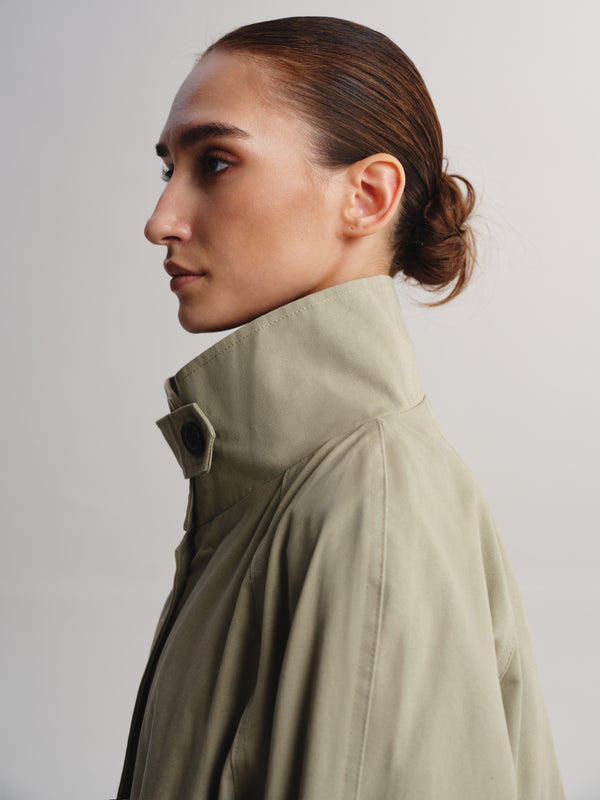 A side-profile portrait of a model wearing a high-neck, light olive green jacket against a plain light gray background.