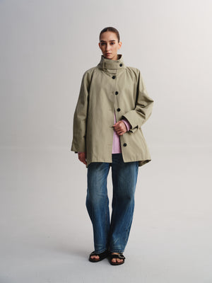 A model stands against a light gray studio background wearing a light olive green high-neck jacket over a pink top, blue jeans, and black sandals.