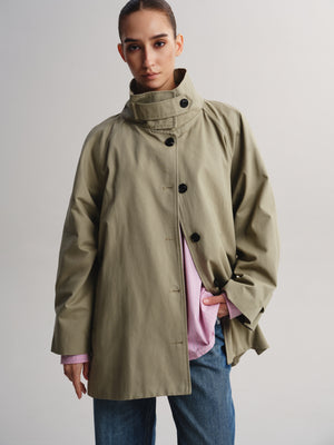 A model wearing a light olive green high-neck coat over a pink shirt and blue jeans, photographed against a neutral light gray studio background.