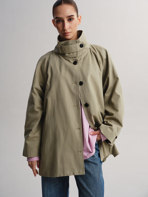 A model wearing a light olive green high-neck coat over a pink shirt and blue jeans, photographed against a neutral light gray studio background.