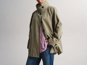 A person wearing an oversized olive green high-collar jacket over a pink shirt and blue jeans, photographed against a plain light gray studio background.