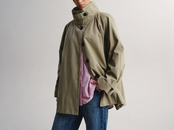 A person wearing an oversized olive green high-collar jacket over a pink shirt and blue jeans, photographed against a plain light gray studio background.