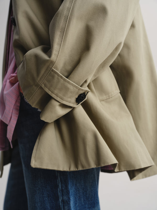 A close-up of a person wearing a light khaki utility jacket with rolled sleeves, a pink shirt underneath, and blue jeans against a plain light background.
