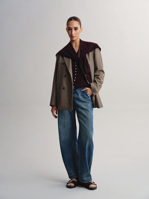 A model stands against a light gray studio background wearing a brown double-breasted jacket with a dark burgundy velvet collar, a dark top, wide-leg blue jeans, and brown sandals.