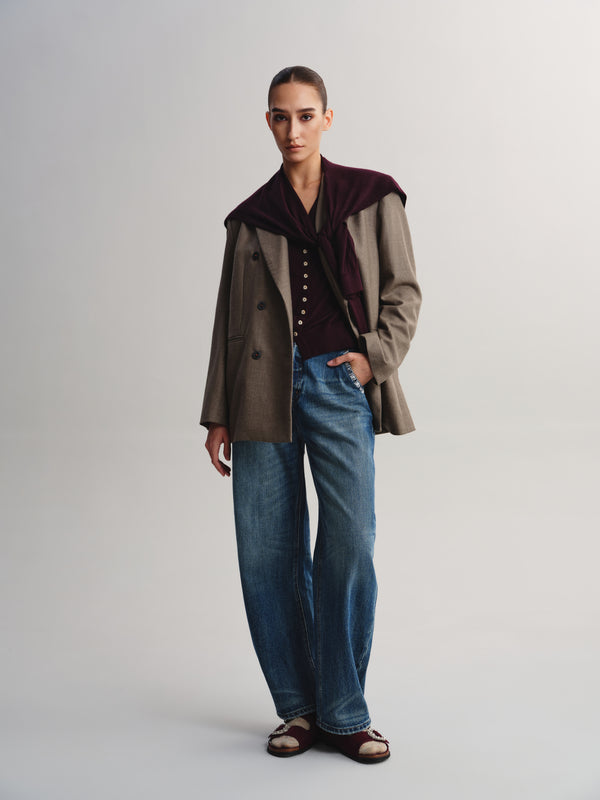 A model stands against a light gray studio background wearing a brown double-breasted jacket with a dark burgundy velvet collar, a dark top, wide-leg blue jeans, and brown sandals.