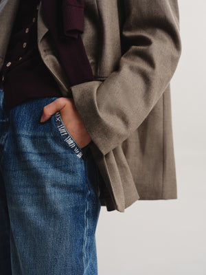 A close-up of a person wearing a taupe-brown textured blazer over a dark burgundy sweater and blue jeans, with one hand in the pocket, against a plain light gray background.
