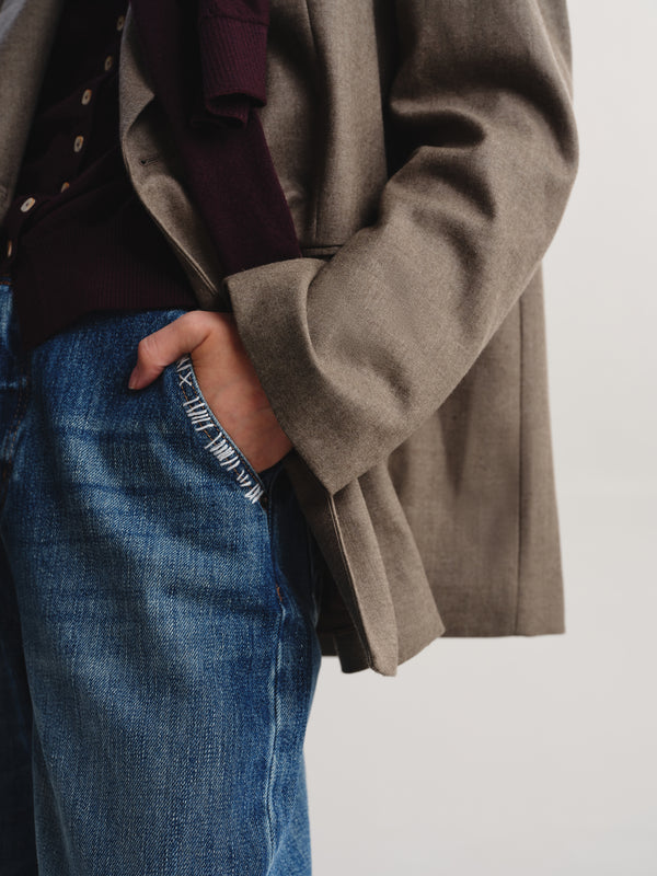 A close-up of a person wearing a taupe-brown textured blazer over a dark burgundy sweater and blue jeans, with one hand in the pocket, against a plain light gray background.