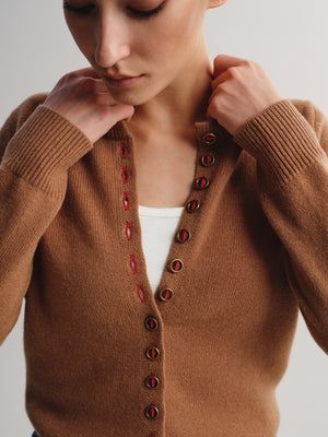 A close-up of a woman wearing a brown knit cardigan sweater with red and gold buttons, layered over a white top, against a light gray background.