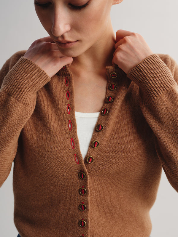 A close-up of a woman wearing a brown knit cardigan sweater with red and gold buttons, layered over a white top, against a light gray background.