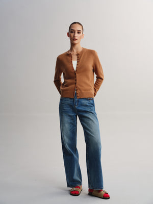 A model stands against a light gray studio background wearing a camel brown cardigan, blue wide-leg jeans, and red-and-beige flat sandals.