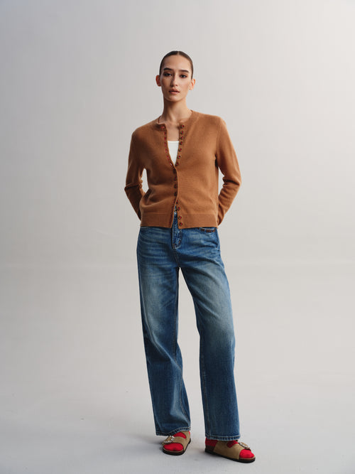 A model stands against a light gray studio background wearing a camel brown cardigan, blue wide-leg jeans, and red-and-beige flat sandals.