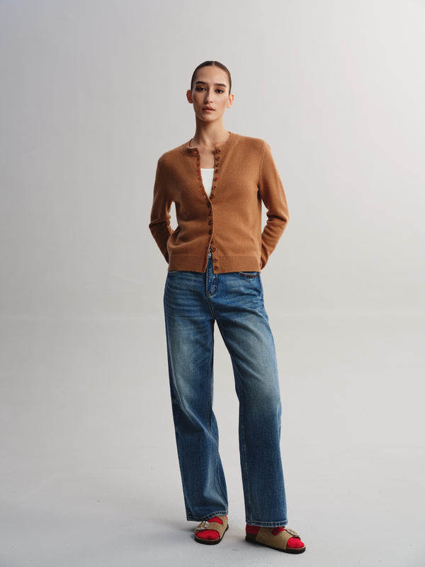 A model stands against a light gray studio background wearing a camel brown cardigan, blue wide-leg jeans, and red-and-beige flat sandals.
