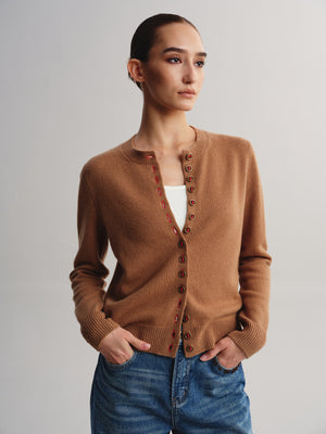 A woman modeling a tan knit cardigan with brown buttons, worn over a white top and blue jeans, against a light gray studio background.