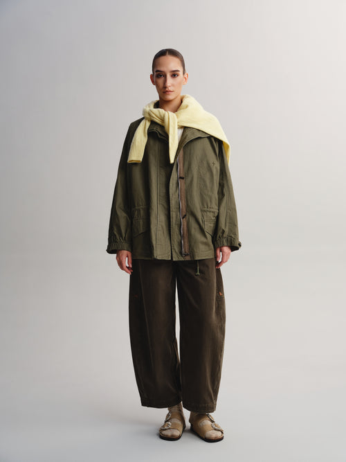 A model stands against a light gray studio background wearing an olive green utility jacket with a pale yellow knit collar, dark brown wide-leg pants, and beige sandals.