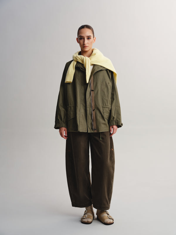 A model stands against a light gray studio background wearing an olive green utility jacket with a pale yellow knit collar, dark brown wide-leg pants, and beige sandals.