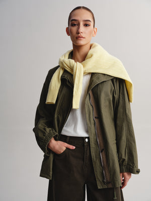A model wearing an olive green utility jacket over a white t-shirt, styled with a thick pale yellow knit scarf draped around the neck and dark brown trousers, shot against a neutral gray studio background.