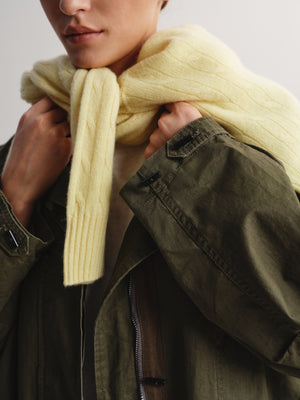 A close-up of a person wearing a pale yellow cable-knit scarf wrapped around their neck, paired with an olive green jacket over a light beige shirt, against a neutral light gray background.