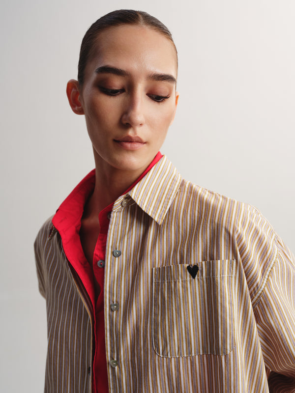 A close-up of a model wearing a beige and brown vertically striped button-down shirt with a red inner collar lining and a small black heart detail on the chest pocket.