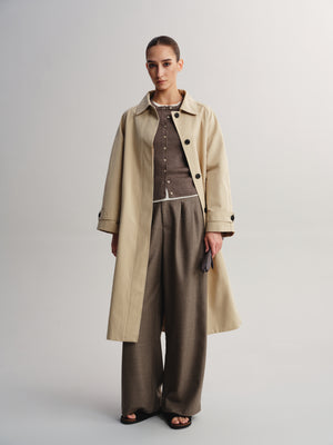 A model stands against a light gray studio background wearing a long beige trench coat over a brown button-front top and wide-leg taupe trousers, with black sandals.