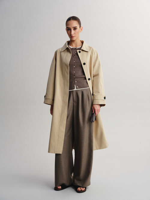 A model stands against a light gray studio background wearing a long beige trench coat over a brown button-front top and wide-leg taupe trousers, with black sandals.