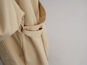Close-up of a beige trench coat with a matching fabric belt tied around the waist, shown against a plain light gray background.