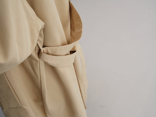 Close-up of a beige trench coat with a matching fabric belt tied around the waist, shown against a plain light gray background.