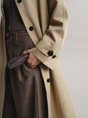 A close-up of a person wearing a beige trench coat over dark brown tailored trousers and a matching belt, with a hand in the coat pocket holding a small dark clutch.