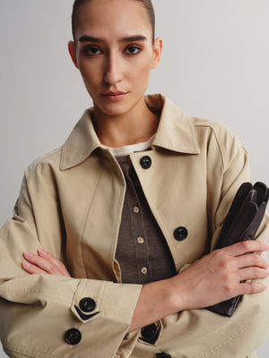 A model with a shaved head wearing a beige trench coat over a brown button-up shirt, holding a dark brown clutch against a light gray studio background.