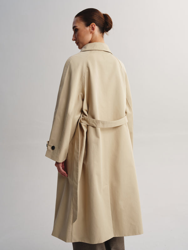 A woman shown from the back wearing a long beige trench coat with a matching waist tie, photographed against a plain light gray studio background.