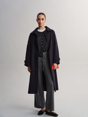 A model stands against a light gray studio background wearing a long dark navy coat over a dark gray button-front top, wide-leg gray trousers, and black flats, while holding a small bright red clutch.