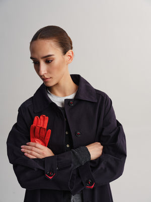A woman wearing a dark navy coat over a white shirt, holding a bright red glove in front of her chest against a light gray studio background.