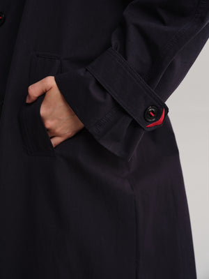 Close-up of a dark navy coat sleeve with a buttoned cuff and a small red accent visible beneath the button, worn by a model whose hand is tucked into a pocket.