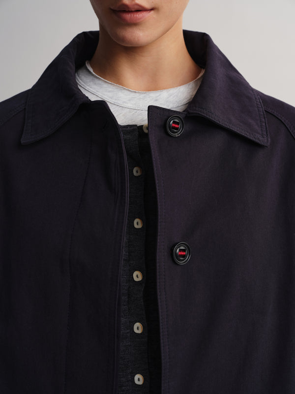 Close-up of a person wearing a dark navy overshirt or jacket over a dark shirt and light gray T-shirt. The collar and front buttons are visible, with small red-and-white logo details on the buttons.