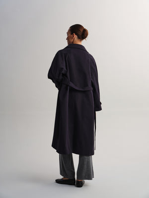 A woman is shown from the back wearing a long dark navy trench coat, gray wide-leg trousers, and black flats against a plain light gray studio background.