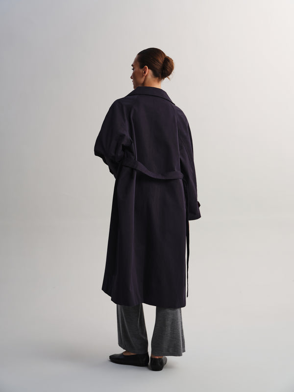 A woman is shown from the back wearing a long dark navy trench coat, gray wide-leg trousers, and black flats against a plain light gray studio background.