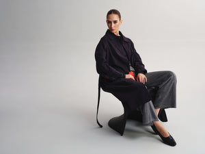 A model sits on a sculptural black chair against a light gray studio background, wearing a dark navy longline jacket, wide-leg charcoal gray trousers, and black pointed flats.