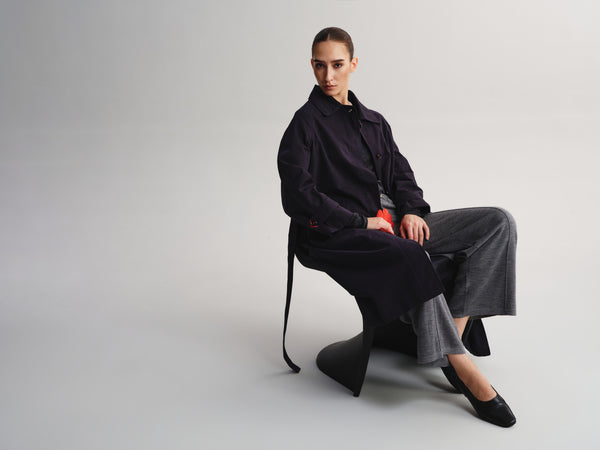 A model sits on a sculptural black chair against a light gray studio background, wearing a dark navy longline jacket, wide-leg charcoal gray trousers, and black pointed flats.