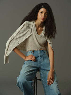 Woman wearing a khaki sweater and blue jeans sitting on a stool against a gray background