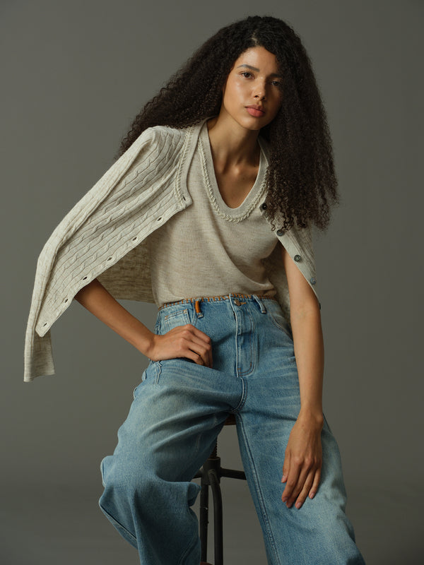 Woman wearing a khaki sweater and blue jeans sitting on a stool against a gray background