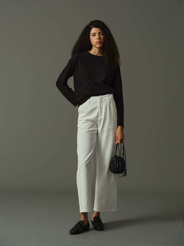 Woman wearing a black sweater and white pants holding a black handbag against a gray background