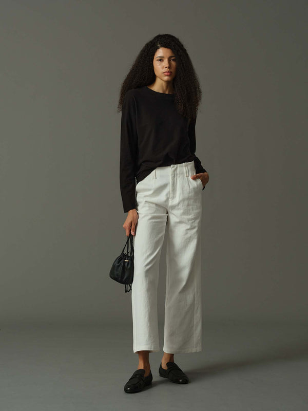 Woman wearing a black top and white pants holding a black handbag against a gray background