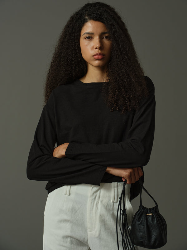 Woman wearing a black long-sleeve top and light-colored pants, holding a black handbag against a gray background.
