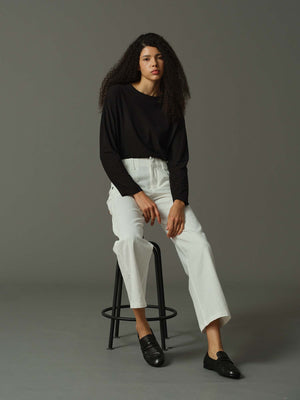 Woman sitting on a stool wearing a black top and white pants against a gray background